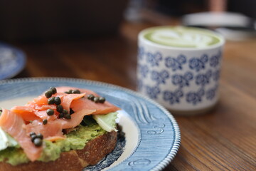 Healthy breakfast with smoked salmon avocado toast topped with capers, served on artisan bread with a cup of matcha latte in a floral ceramic mug. Cozy café setting with rustic wooden table.