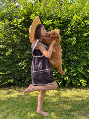 Young girl in summer dress and straw hat joyfully holds fluffy dog in a lush green garden,...