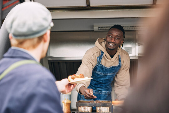 Young man wearing apron serving hot dog to customer while standing in food truck