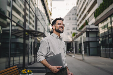 one man with beard and eyeglasses walk with suitcase and hold laptop