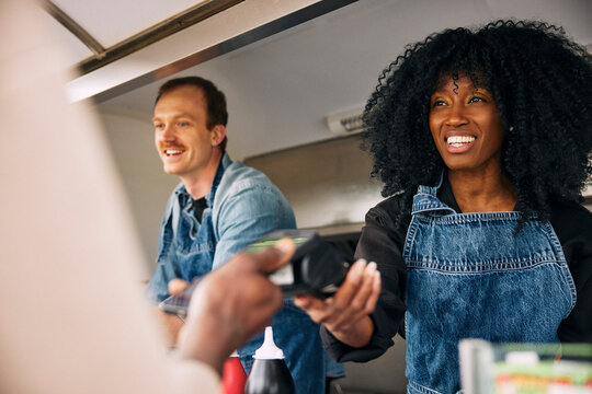 Smiling curly haired female owner holding credit card reader for woman making payment at food truck
