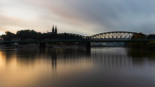 Historic railway bridge and Vysehrad silhouette reflected in the calm Vltava River before sunrise in Prague, Czech Republic. Atmospheric long exposure cityscape with soft morning light.