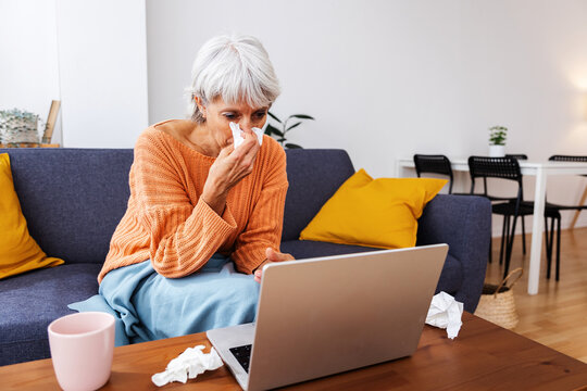 Senior sick woman wrapped in a warm blanket on sofa blowing her nose while talking with doctor through virtual meeting on video call from home