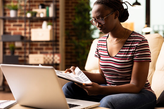 Determined female entrepreneur carefully reviewing printed documents during remote work session. Focused black woman flips through clipboard, preparing notes for upcoming project presentation.