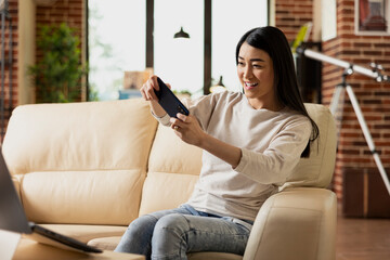 Obraz premium Confident self employed woman relaxes on sofa while playing video game on smartphone. Asian female entrepreneur with mobile device, enjoying fun break from work in cozy brick wall living room.
