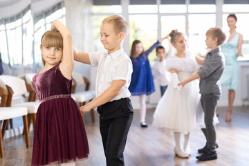 Smiling junior schoolchildren in party dresses learning dancing waltz with pedagogue in social hall