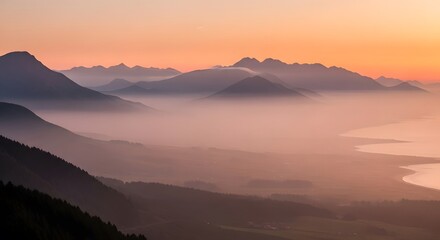 Panoramic view of mountain range enveloped in mist at sunset. Orange sky over distant peaks reflects in calm lake below. Forested slopes lead to valley with town.