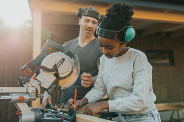 Girl wearing noise cancellation headphones while marking measurement on wooden plank with help of father outside house