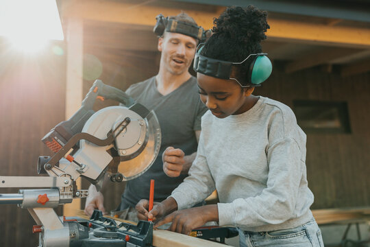 Girl wearing noise cancellation headphones while marking measurement on wooden plank with help of father outside house