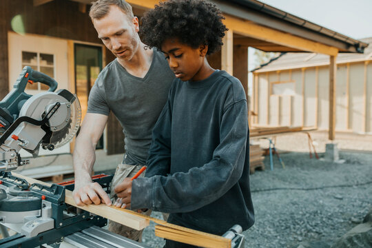Curly haired boy marking measurement on wooden plank with help of father outside house