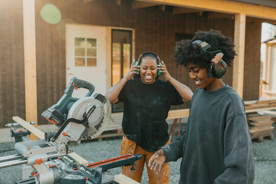 Happy mother looking at son wearing noise cancellation headphones cutting wooden plank with circular saw in back yard