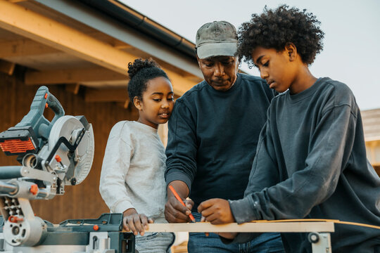 Grandfather teaching grandchildren to mark measurement on wooden plank for home renovation
