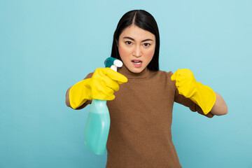 Annoyed asian housemaid in rubber gloves holding detergent spray in aggressive pose. Angry female...