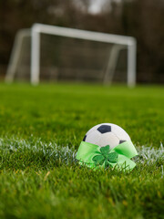 Obraz premium Green bow with Irish shamrock by a classic football or soccer ball on a green grass training field. Goal post in the background. Supporting national team of Ireland concept.