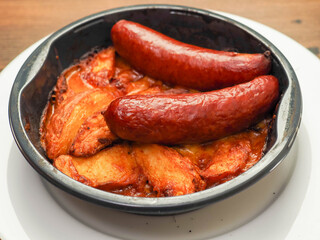 Cooked dish with potato wedges, sauce, cheese and two pork sausages in a plastic dish. Ready made meal made from high quality products. Tasty dinner on a wooden table.