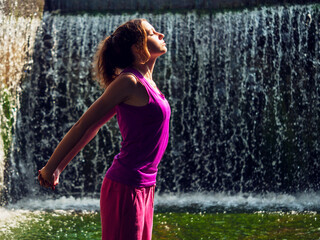 Young fitness girl model is standing in river water with stunning waterfall background. Girl is...