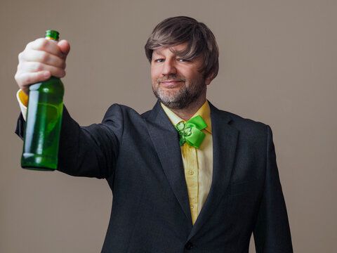 Man in grey suit and yellow shirt and green bow tie with shamrock is holding green bottle of lager beer in his hand. Irish sport fan and supporter concept. Football and rugby fan. Stylish male model