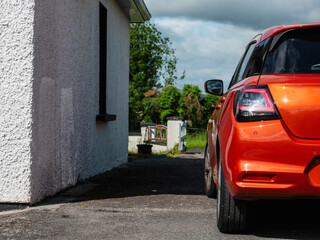 Small sport red car parked by a house with white walls, rural area in the background. Warm sunny...