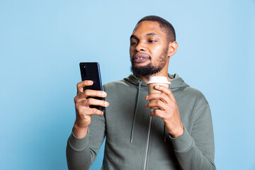 Positive friendly guy drinks coffee and checks his smartphone, answering to online messages being...
