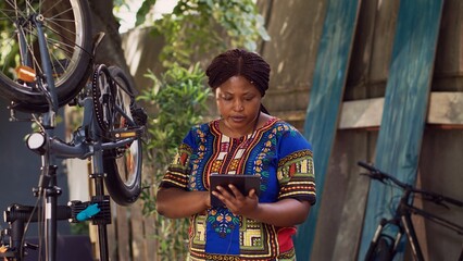 Detailed view of sports-loving black woman searching on phone tablet for bike summer maintainance. African american female grasping smart device while inspecting and maintaining bicycle.
