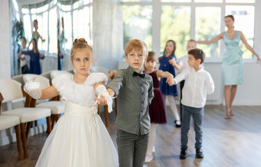Enthusiastic preteen children, boys and girls in party attire performing elegant waltz in pairs in sunny hall of dance school with smiling female teacher in background..