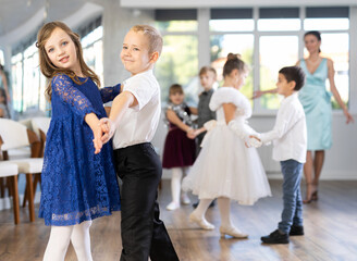 Naklejka premium Happy little children in elegant dresses practicing waltz dance with teacher in school hall