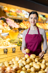 Pleased middle-aged saleswoman placing potatoes on counter in large grocery store