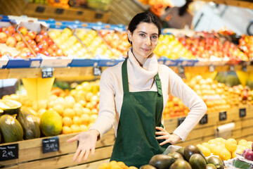 Young woman seller in apron offers fresh avocado in vegetable shop