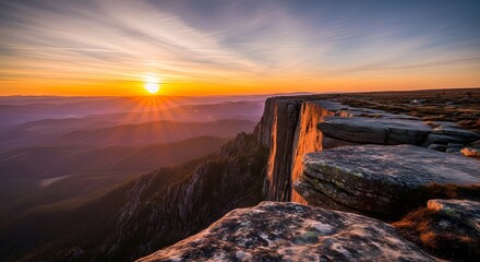 Sunset illuminates a rugged cliff edge showcasing textured rocks against a backdrop of layered mountain ranges and a vibrant sky