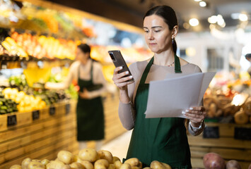 Woman in an apron works in the vegetable department, checks papers about the presence of vegetables and fruits and holds a phone in her hands