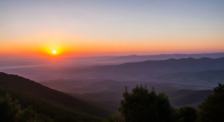 Obraz premium A vibrant sunset unfolds over a mountainous landscape with a clear sky transitioning from orange to blue. Rolling hills create a layered effect leading to the horizon with the sun.