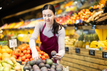 Positive middle-aged female seller putting avocados on stand in fruit and vegetable market