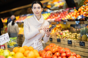 Female looking at shop windows in greengrocery with phone, planning to buy ripe fruit and vegetable. Client scrolls through list of reminders and notes on phone.
