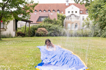 Child enjoying summer fun on a colorful water slide in a lush green backyard, surrounded by playful...