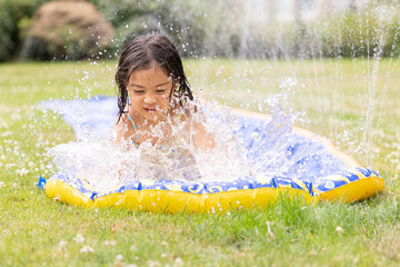 Child joyfully splashes water while sliding on inflatable mat in sunny garden, capturing the...