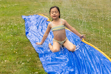 Child enjoying summer fun on a water slide, splashing in the air, surrounded by green grass and a...