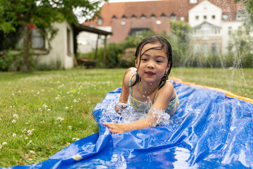 Child enjoys summer fun sliding on a blue tarp with water splashing around, surrounded by green...