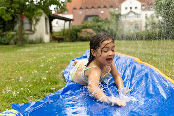 Child enjoying summer water activities, sliding on a blue water slide in a garden, surrounded by...