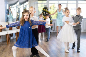 Smiling junior schoolchildren in party dresses learning waltz with pedagogue in hall embellished...