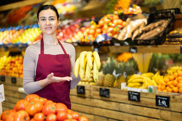 Polite middle-aged female seller offering bananas standing by counter in large grocery store