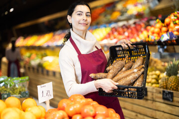 Young woman seller in apron offers cassava in box in vegetable shop