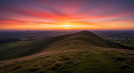 Scenic landscape photograph featuring a vibrant sunset over a rolling green hill with a distant town and colorful sky