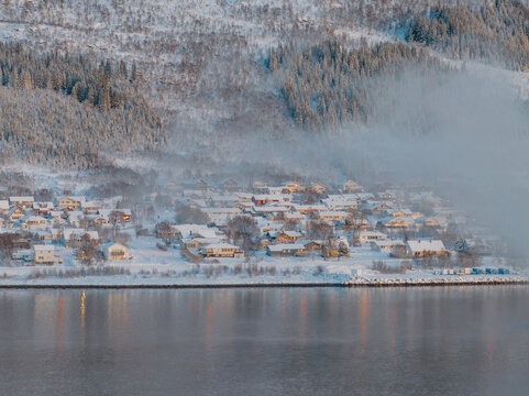 Snowy village at fjord&rsquo;s edge with rising mist