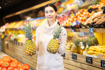 Woman chooses ripe pineapples against the background of shelves with vegetables and fruits in a supermarket