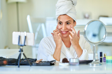 Woman in bathrobe putting cream on her face in front of mobile phone.