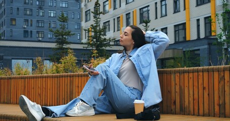 Young woman wearing casual clothes sits on a wooden bench, sipping coffee from a disposable cup and...
