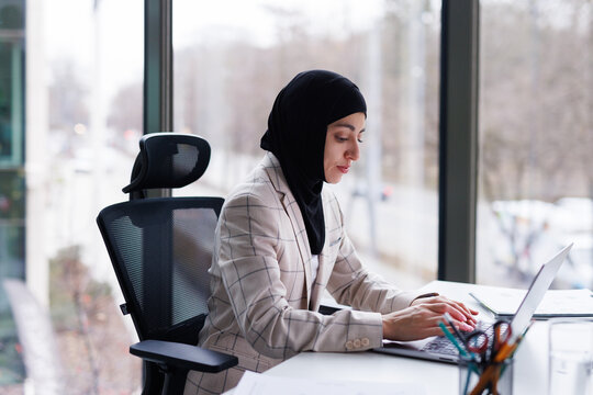 CEO in hijab typing text on laptop sitting at desk near window