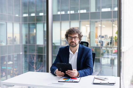 CEO in business suit sitting at desk in office with tablet in hands 