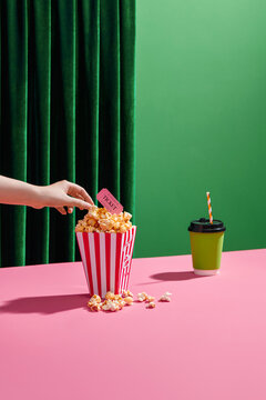 Hand Holding Movie Ticket Over Popcorn with Drink on Pink Table
