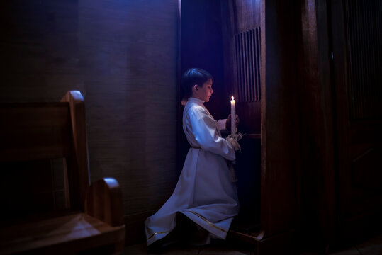 Catholic boy holding a candle, confessing before his first communion. Child in white clothes kneeling in the dark in front of the confessional.